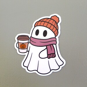 May include: A white ghost sticker wearing an orange beanie and a pink scarf, holding a coffee cup with a pumpkin design. The coffee cup has a brown lid and a white body. The ghost has black eyes.