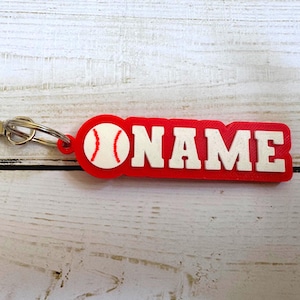 May include: A red and white keychain featuring a baseball graphic on the left side and the word NAME in bold, raised white capital letters on a red rectangular base, attached to a silver-colored metal lobster clasp and a circular split ring.