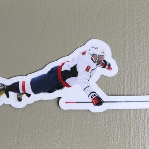 May include: A hockey player wearing a white and red jersey with the number 8 on it, is sliding on the ice with a hockey stick in hand. The player is wearing a white helmet with a black visor.