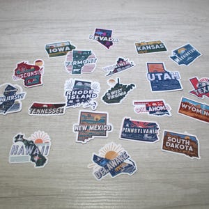 May include: A collection of 20 colorful state-shaped stickers with names and illustrations. The stickers are on a light brown wooden surface.