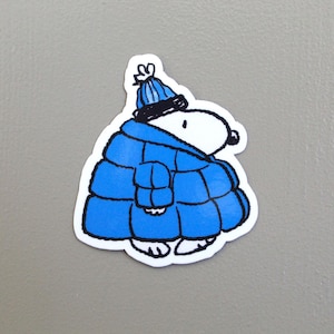 May include: A sticker featuring a cartoon character, Snoopy, dressed in winter attire. Snoopy is wearing a blue quilted jacket and a matching blue hat with a black brim. The sticker has a white border and is set against a gray background.