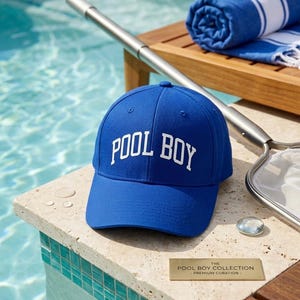 May include: A royal blue baseball cap featuring the white embroidered text POOL BOY on the front panel. The cap has a structured crown, a curved brim, and eyelets for ventilation. A small rectangular gold plaque nearby reads THE POOL BOY COLLECTION.