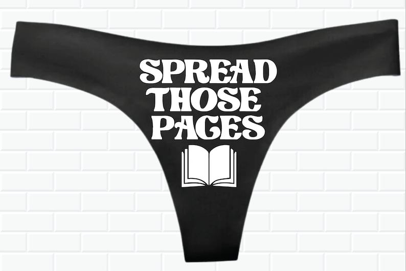 May include: Black thong underwear with white text that reads "SPREAD THOSE PAGES" and a white outline of an open book.