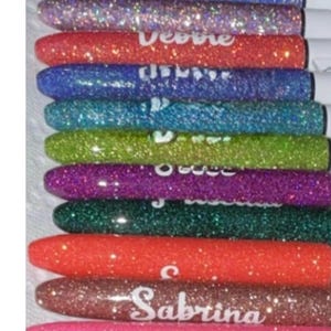 Puede incluir: Una pila de bolígrafos con purpurina de varios colores, cada uno con un nombre escrito. Los bolígrafos son rosas, azules, morados, rojos, verdes y dorados. Los nombres incluyen Kelsey, Jam, Debbie y Sabrina.