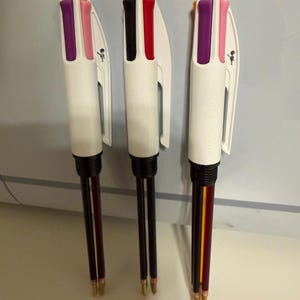 以下が含まれることがあります： 白と複数の色のインクが入った3本のマルチカラーペン。各ペンには、紫、ピンク、黒、赤、黄色、オレンジの異なるインク色の組み合わせがあります。ペンには、ストラップを取り付けるための小さなループが上部にあります。
