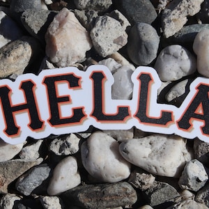 May include: A white sticker with the word "HELLA" in black letters with an orange outline. The sticker is on a bed of small gray rocks.