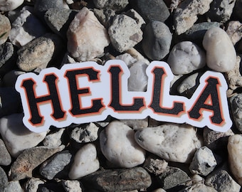 May include: A white sticker with the word "HELLA" in black letters with an orange outline. The sticker is on a bed of small gray rocks.