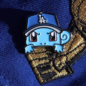 Puede incluir: Un pin de esmalte azul y blanco de un personaje de dibujos animados que lleva una gorra de béisbol con las letras "LA" en ella. El personaje tiene la piel azul, los ojos rojos y una boca blanca. El pin está sujeto a un fondo de tela dorado y negro.