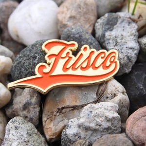 May include: A Frisco pin with a gold border and a red and cream-colored design. The pin is in the shape of a wordmark and is set against a background of various-sized rocks and pebbles.