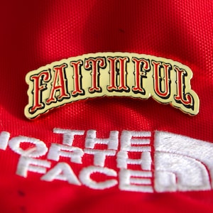 Peut inclure: Épinglette dorée avec le mot "FAITHFUL" en rouge, souligné de noir, arqué vers le haut. L'épinglette est posée sur un fond rouge avec le logo blanc brodé de "THE NORTH FACE".