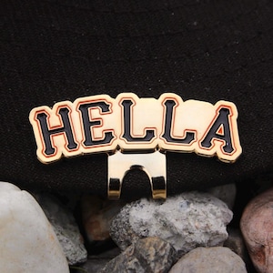 May include: A gold-coloured hat clip with the word "HELLA" in black letters with a red outline. The clip is attached to a black hat.