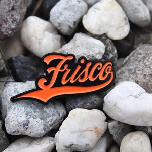 May include: A black and orange Frisco patch with a stylized script font, resting on a bed of gray and white rocks. The patch has a black border and a bright orange interior.