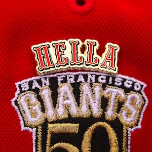 May include: A red baseball cap with a gold embroidered patch that says "San Francisco Giants" with a gold "50" in the center. Above the patch is a gold pin that says "HELLA" in red letters.