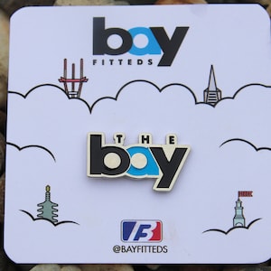 May include: A white card with black outlines of San Francisco landmarks. The card features a black and blue enamel pin with the text "THE BAY" in white letters. The top of the card has the text "bay FITTEDS" in black letters. The bottom of the card has the text "@BAYFITTEDS" in black letters.