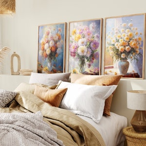 Op de afbeelding: Drie ingelijste bloemschilderijen met boeketten van roze, witte en gele bloemen in vazen. De schilderijen hangen aan een witte muur boven een bed met bruin en wit beddengoed.
