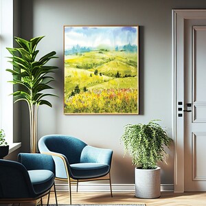 Puede incluir: Una pintura de acuarela de una colina verde ondulada con un cielo azul y nubes blancas en el fondo. La pintura está enmarcada en un marco de madera claro y está colgada en una pared en una habitación con una planta verde y dos sillas azules.