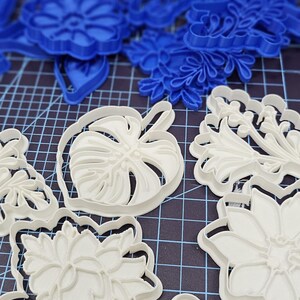 May include: An assortment of plastic floral and leaf-shaped cookie cutters in white, blue, and black. The cutters are arranged on a dark surface with a grid pattern, ideal for baking and crafting.