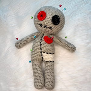May include: A handmade gray crochet voodoo doll with a red heart, a red button eye, and a black button eye. The doll has black stitched details and is adorned with colorful pins. The doll is lying on a white fluffy surface.