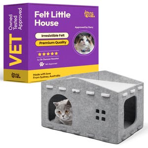 Op de afbeelding: Een grijze vilten kattenhuis met een wit dak en een kat die uit de ronde deuropening kijkt. Het huis heeft een vierkant raam en witte decoratieve accenten. Een paarse doos met de tekst "Felt Little House" en "Vet Approved" staat op de achtergrond.