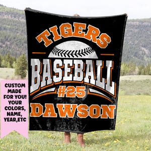 May include: Black baseball blanket featuring the text "TIGERS BASEBALL #25 DAWSON" in white and orange. A baseball graphic is centered above the word "BASEBALL". The blanket is displayed against a backdrop of a grassy field and mountains.