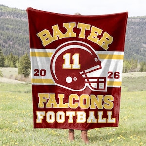 Pode incluir: Um cobertor bordô e dourado com tema de futebol. O cobertor apresenta as palavras "BAXTER FALCONS FOOTBALL" em letras douradas, juntamente com um gráfico de capacete de futebol e os números "11", "20" e "26".