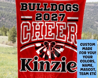 May include: A red blanket with the words "BULLDOGS 2027 CHEER Kinzie" in white and black lettering. A bow graphic is centered below "CHEER". A blue rectangle on the right says "CUSTOM MADE FOR YOU! YOUR COLORS, NAME, MASCOT, TEAM ETC".