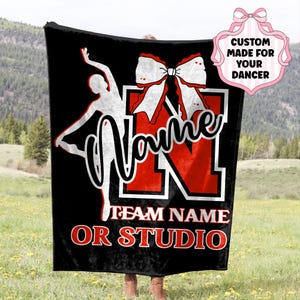 May include: Black blanket with a red and white design. Features a dancer silhouette, the word "Name" in script, and a large "N" with a bow. Text at the bottom reads "Team Name or Studio". The blanket is custom made for a dancer.