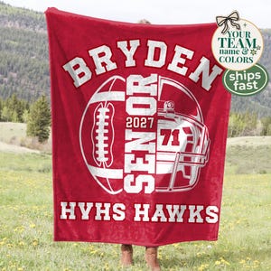 May include: A red fleece blanket featuring a white graphic of a football and a helmet. The text reads BRYDEN, SENIOR, 2027, 71, and HYHS HAWKS in bold white lettering. The design is centred on the fabric, showcasing a sports-themed layout.