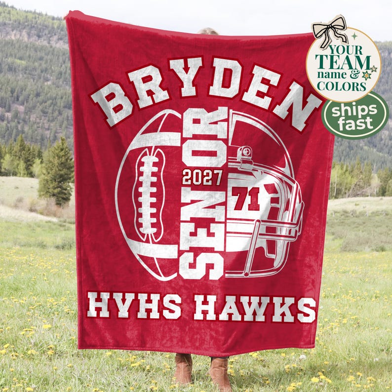 May include: A red fleece blanket featuring a white graphic of a football and a helmet. The text reads BRYDEN, SENIOR, 2027, 71, and HYHS HAWKS in bold white lettering. The design is centred on the fabric, showcasing a sports-themed layout.