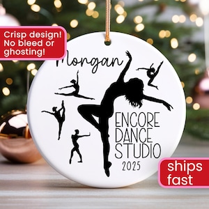 May include: A white ceramic ornament with a gold string for hanging. The ornament features a black silhouette of a dancer and the text "ENCORE DANCE STUDIO 2025". The name "Morgan" is written above the dancer.