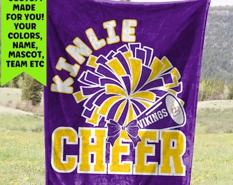 May include: A purple blanket with a cheerleading theme. The blanket features the name "Kinlie" in white letters above a pom-pom design in purple and gold. The word "CHEER" is in large gold letters.