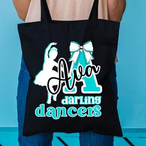 May include: Black tote bag with a white ballerina silhouette and the name "Ava" in a script font, with the words "darling dancers" in a teal font. A teal bow is also featured on the bag.