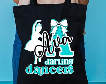 May include: Black tote bag with a white ballerina silhouette and the name "Ava" in a script font, with the words "darling dancers" in a teal font. A teal bow is also featured on the bag.