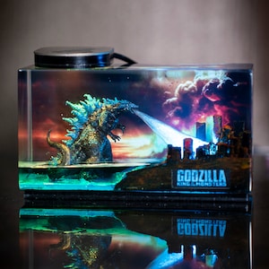 Könnte beinhalten: Miniatur-Diorama mit Godzilla, der einen Energiestrahl speit, vor einer Stadtlandschaft und einem dramatischen Himmel. Auf der Basis des Dioramas steht der Text "GODZILLA KING OF THE MONSTERS". Die Szene ist in einem transparenten, rechteckigen Block eingeschlossen.