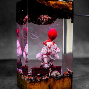 Op de afbeelding: Een rechthoekig harskunstwerk met een clownfiguur die een rode ballon vasthoudt. De clown draagt een wit en rood kostuum. Het blok heeft een donkere bovenkant en een houten basis, met schedels en andere figuren zichtbaar.