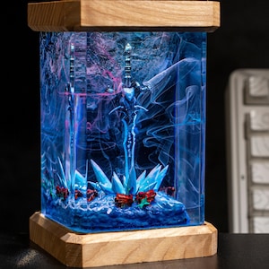 May include: A rectangular resin art piece featuring a sword design encased in blue resin with red and blue accents. The piece is set on a wooden base and topped with a wooden frame.