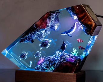 May include: A diamond-shaped resin lamp featuring an underwater scene with miniature sea turtles, a whale, a manta ray, and clownfish. The clear resin is embedded with dark wood elements, coral-like structures, and internal LED lighting.