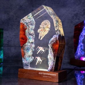 Può includere: Una lampada in resina con una base trasparente e un piedistallo in legno. La resina contiene un modello in miniatura della nave spaziale Millennium Falcon di Star Wars. La lampada è illuminata dal basso, illuminando la resina e la nave spaziale.