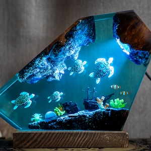 May include: A resin lamp shaped like a diamond with a blue and green ocean scene inside. The lamp features a wooden base and a light that illuminates the scene, which includes a shipwreck, coral, and several sea turtles.