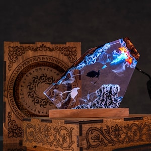 May include: A unique resin art piece featuring a layered design with blue, white, and orange hues, depicting an underwater scene with silhouettes of marine life. The art piece is mounted on a wooden base and sits in front of a wooden box.