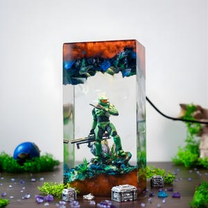 Puede incluir: Lámpara rectangular de resina con una figura de acción verde sosteniendo un rifle, sobre un fondo de tonos azules y naranjas. La lámpara tiene un cuerpo transparente con base y tapa de madera. La lámpara mide unos 20 cm de altura.