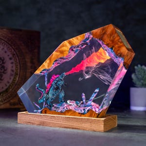 Könnte beinhalten: Dekorative Resin-Skulptur mit einer blau-rosa Godzilla-Figur, die Feuer speit. Die Skulptur ist in klarem Harz mit Holzakzenten eingeschlossen und steht auf einem Holzsockel. Das Feuer ist eine Mischung aus Rot, Orange und Pink.