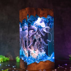 May include: A rectangular wooden lamp with a detailed resin design. The lamp features a warrior figure encased in blue resin with a smoky effect. The top and bottom are made of wood, and the lamp emits a soft, ambient glow.