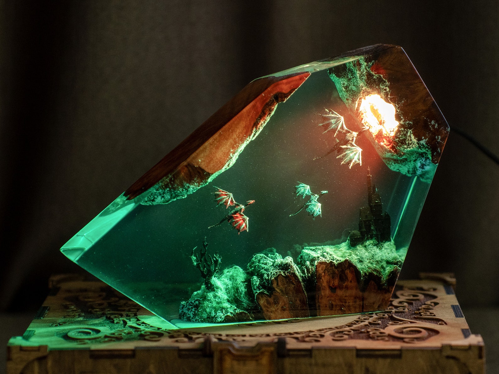 In an unusual geometric shape, three dragons soar through a resin sculpture with a ground appearing to be made of stone housing a castle and a gnarled tree, towards a glowing circular light. The sculpture rests on an engraved wooden box-shaped stand.