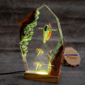 May include: A unique resin lamp featuring a spaceship and two smaller aircraft, set against a backdrop of brown and green rock formations. The lamp has a wooden base and a clear, geometric frame. The design includes a warm, yellow light.