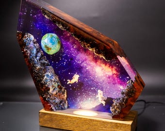 Può includere: Una lampada in resina con un design a tema galattico e astronauti. La lampada ha una forma triangolare e una base in legno. La galassia è di un viola e rosa vibrante con un piccolo pianeta blu nell'angolo. Due astronauti galleggiano nella galassia.