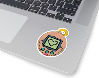 May include: A sticker featuring a retro digital pet design. The sticker has a peach-colored background with a green screen displaying a pixelated heart. The design is framed with blue accents, floral elements, and a yellow loop.