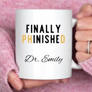 May include: White ceramic mug with the words "FINALLY PHINISHED" in black and gold lettering, with "Dr. Emily" in script below. The mug is held by a person wearing a pink sweater.