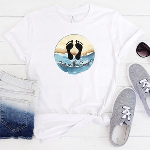 May include: White t-shirt with a circular graphic featuring black footprints, sand, and blue water, with the text "Toes in the Sand". The shirt is paired with denim shorts, gray sneakers, and sunglasses.