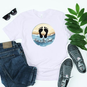 May include: White t-shirt with a circular graphic featuring black footprints on a beach with the text "Toes in the Sand." The shirt is paired with blue jeans, sunglasses, and gray sneakers, suggesting a casual, beach-themed style.